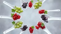 Fresh berries on white spoons arranged in circle pattern with red currants, black currants and gooseberries for culinary presentation and food styling.