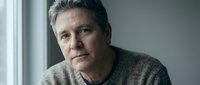 Mature Caucasian man in gray sweater sitting by window with thoughtful expression, natural lighting for lifestyle and wellness concepts.