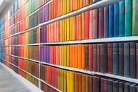 Colorful rainbow arrangement of books on library shelves creating vibrant spectrum pattern, organized by color from red to blue on multiple rows of bookshelves.