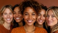 Diverse group of smiling women with different skin tones and hair types posing together against warm brown background, expressing joy and friendship in casual setting.