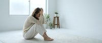 Young woman experiencing depression sitting alone on floor in minimalist room holding head in hands showing mental health struggles and emotional distress.