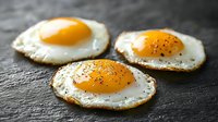 Three sunny side up eggs with black pepper seasoning on dark slate stone background, crispy edges and runny golden yolks for breakfast or brunch menu.