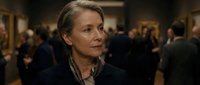 Mature Caucasian businesswoman with blonde hair in elegant black coat at upscale corporate networking event with professionals in background.