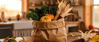 Fresh organic vegetables and corn in brown paper grocery bag on kitchen counter during golden hour sunlight for healthy cooking and meal preparation.