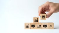 Hand placing wooden cube with skull danger symbol on stack of business icon blocks representing risk management and corporate safety assessment.