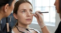 Professional makeup artist applying eyebrow makeup to young Caucasian woman client in modern beauty salon with natural lighting for cosmetic services.