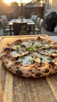 Freshly baked pizza with charred crust and melted cheese on wooden board, outdoor patio dining setting with table and chairs during golden hour sunset.