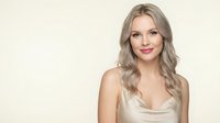 Young blonde woman with wavy hair smiling at camera wearing gold satin top against beige background for beauty and fashion content.