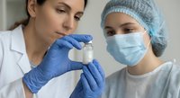 Two female medical professionals examining vaccine vial in laboratory setting wearing protective equipment and surgical masks for healthcare research.