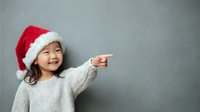 Cheerful Asian girl in Santa hat pointing at copy space on gray wall background, expressing joy during holiday season.