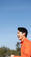 Young Asian male in bright orange shirt laughing outdoors against clear blue sky. Perfect for lifestyle, happiness, and wellness campaigns.