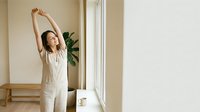 Woman in casual beige linen clothing stretching arms overhead near bright window in minimalist interior space, conveying wellness and morning routine for lifestyle and home decor use.