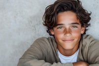 Young Hispanic male teen with wavy dark hair and bright smile wearing casual beige sweater and white t-shirt against textured gray wall background. Genuine expression.