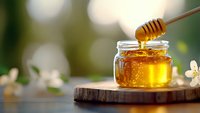 Golden honey dripping from wooden dipper into glass jar on rustic wooden slice against blurred natural background with white spring blossoms and bokeh lights.