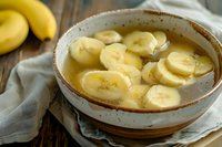 Fresh banana slices floating in sweet honey syrup served in rustic ceramic bowl on wooden table with natural linen napkin and whole bananas in background.