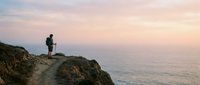 Hiker with backpack standing on rocky mountain cliff overlooking vast ocean horizon at sunset, adventure travel and outdoor exploration concept for lifestyle and wellness projects.