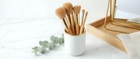 Makeup brushes with gold handles in a white ceramic holder on marble surface, eucalyptus branch and mirror, beauty vanity setup.