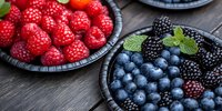 Fresh raspberries, blackberries and blueberries in dark woven bowls garnished with mint leaves on rustic wooden surface, macro food photography.