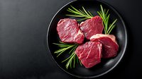 Raw beef tenderloin steaks with fresh rosemary sprigs on black ceramic plate against dark background, fresh premium cut meat for restaurant menu.