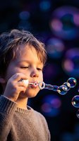Young Caucasian boy blowing soap bubbles with colorful bokeh lights in background creating magical childhood moment for family and lifestyle concepts.