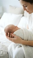 Young Caucasian mother in white gently holds sleeping newborn wrapped in knit blanket, gazing with tender love in soft bright bedroom.