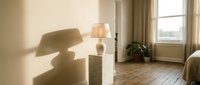 Sunlit modern bedroom interior with wooden floor, table lamp casting shadow on beige wall, draped curtains, potted plant, and minimalist decor for home design projects.
