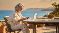 Senior woman with gray curly hair working on laptop at seaside terrace during golden hour, enjoying remote work lifestyle by Mediterranean coast.