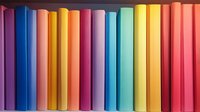 Colorful book spines arranged in rainbow order on bookshelf, creating vibrant spectrum pattern. Clean minimal composition shows organized row of hardcover books.