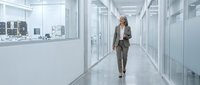 Businesswoman in formal attire walking through modern office corridor with glass partitions and city skyline view, suitable for corporate and professional concepts.