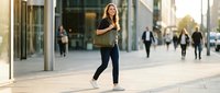 Young professional woman walking confidently through modern business district during golden hour, carrying laptop bag for corporate lifestyle content.