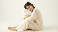 Young woman with dark hair and East Asian ethnicity, wearing cream-colored knit sweater, sitting on floor hugging knees in relaxed pose against white background for wellness and lifestyle content.