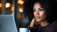 Young African American woman working on laptop in cozy cafe with warm lighting, thoughtful expression, remote work and digital lifestyle concept.