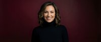 Professional middle-aged Caucasian businesswoman in black turtleneck sweater smiling confidently against dark burgundy background for corporate headshots.