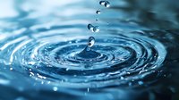 Water drop falling into calm blue water creating concentric ripples and splash with floating droplets captured in high speed macro photography.