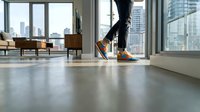 Person wearing orange and black sneakers walking on polished concrete floor in modern loft apartment with floor-to-ceiling windows overlooking city skyline and contemporary furniture.