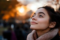 Young Asian woman with gentle smile looking up at sunset, wearing cozy scarf. Natural golden hour lighting creates warm atmospheric portrait with bokeh background.