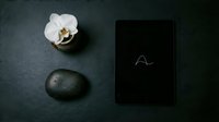 Dark slate surface with tablet, smooth stone, and white orchid in minimalist zen composition for wellness, spa, or tech branding.