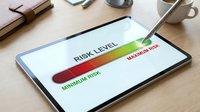 Digital tablet displaying colorful risk assessment gauge from minimum to maximum levels on wooden desk with business accessories for financial analysis.