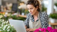 Focused female florist in checkered shirt and apron working on laptop among colorful flowers in modern flower shop, managing online orders.