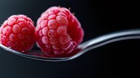 Fresh ripe raspberries on silver spoon against dark background, macro food photography showing texture and details of juicy red berries.