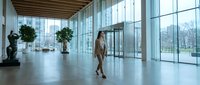 Professional businesswoman walking through modern corporate office lobby with floor-to-ceiling windows and natural lighting for business concepts.