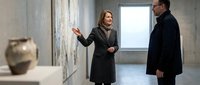 Professional Caucasian female real estate agent in dark coat presenting modern apartment interior to male client during property viewing consultation.