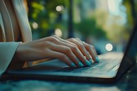 Female hands with turquoise manicure typing on laptop keyboard in soft evening light with bokeh effect background, showing modern workplace lifestyle.