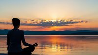 Young woman practicing yoga meditation in lotus pose by tranquil lake at golden sunset with peaceful sky reflection creating serene wellness atmosphere.