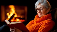 Senior Caucasian woman in orange knit sweater reading book by fireplace, smiling contentedly. Cozy winter evening atmosphere with warm fire glow creates relaxing home scene.