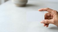 Hand holding blank white card on neutral surface with blurred background, suitable for business presentations, marketing mockups, and promotional designs.