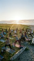 Diverse group practicing yoga and stretching exercises on beach grass at sunset, including person in wheelchair, promoting inclusive fitness and wellness outdoors.