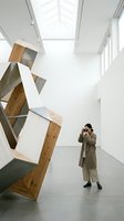 Woman in beige coat photographing abstract wooden and metal sculpture in minimalist gallery space with skylights and polished concrete floors.