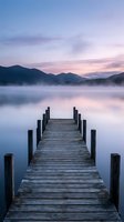 Wooden jetty extending into misty lake at dawn with mountain silhouettes and pastel sky reflections creating peaceful morning atmosphere for meditation and relaxation.