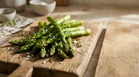 Fresh green asparagus spears seasoned with herbs on rustic wooden cutting board in bright kitchen setting for healthy cooking and meal preparation.
